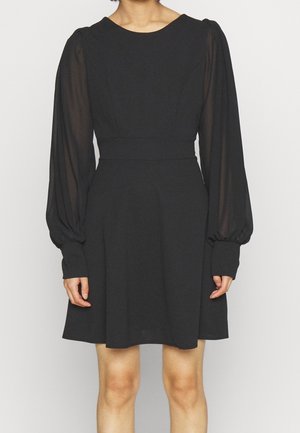 Donna che indossa un vestito nero con maniche lunghe a sbuffo trasparenti, vita aderente e gonna svasata fino al ginocchio, in piedi davanti a uno sfondo neutro.