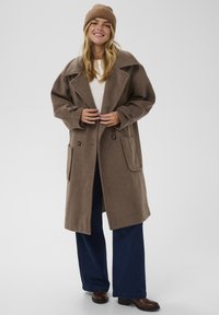 Hnědý vlněný kabát s širokým límcem, dvojitým zapínáním na knoflíky a velkými předními kapsami, nošený přes krémový top a tmavě modré kalhoty.