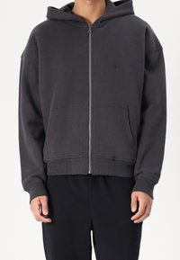 Donkergrijze zip-up hoodie gemaakt van een zacht materiaal. Beschikt over een kangoeroezak en een capuchon met koord. Geribde manchetten en onderkant.