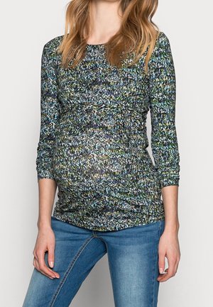 Zwangere vrouw die een meerkleurige, met patronen bedrukte top met lange mouwen en een blauwe spijkerbroek draagt, staand met één hand rustend nabij haar dij.