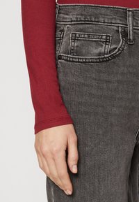 Jeans de mezclilla negra con una textura desgastada, con una cintura con botones y un bolsillo lateral, combinados con una camiseta roja de manga larga.