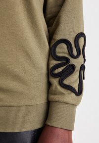 Bluza w kolorze oliwkowej zieleni z prążkowanymi mankietami, z czarnym, teksturowanym haftem kwiatowym na lewym rękawie, przedstawiająca gładką tkaninę.