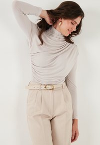 Lichtbeige lange mouwen top met gedrapeerde stof, gecombineerd met hooggotige beige broek die vastzit met een brede riem met een gouden gesp.
