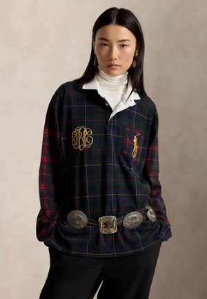 Femme portant une chemise à carreaux sombre avec un col roulé blanc, un pantalon noir à jambes larges, une ceinture décorative et des chaussures noires, debout devant un fond beige uni.
