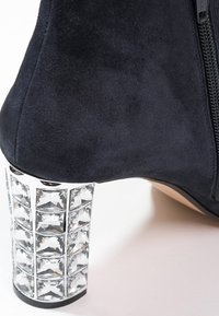 Bota de tobillo de ante azul marino con acabado mate, presentando un tacón bloque adornado con cristales transparentes dispuestos en un patrón de cubo. Cierre con cremallera en el lateral.