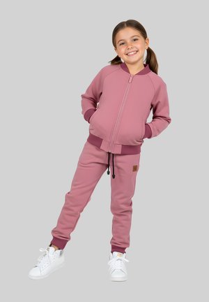 Uśmiechnięta dziewczyna z brązowymi kitkami ubrana w fioletową kurtkę na zamek i pasujące dresy, stoi z rękami w kieszeniach i białymi sportowymi butami.