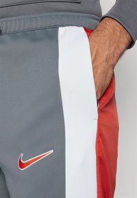 Mano parcialmente en el bolsillo de pantalones de chándal Nike grises, blancos y rojos, con el logo en forma de swoosh rojo de Nike en el muslo.