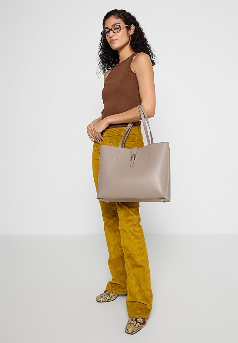 Beige synthetische Tragetasche mit rechteckiger Form, doppelten Griffen und dezenten goldenen Hardware-Akzenten. Strukturierte Stoffhose in senfgelb.