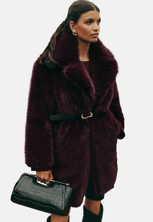 Vínový kožuch z umelého kožucha s širokým golierom, opasok v páse, v kombinácii s lesklou čiernou kabelkou s textúrou krokodílej kože.