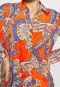 Knapp-up skjorta med en livlig orange bas. Har paisley-mönster i blått, persika och vitt. Tillverkad av ett mjukt tyg med uppvikt ärm.