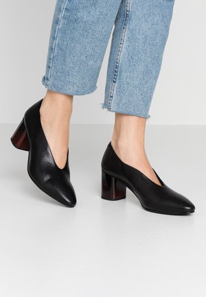 Černé kožené boty na vysokém podpatku s špičatou špičkou a V-výřezem, se silným, lesklým želvovinovým podpatkem. Nosené s světle modrými džíny.