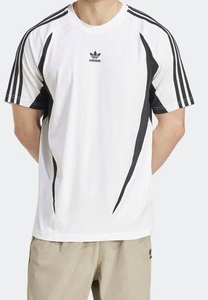 Person iført en hvid Adidas T-shirt med sorte striber på ærmer og sider, kombineret med beige Adidas-shorts.