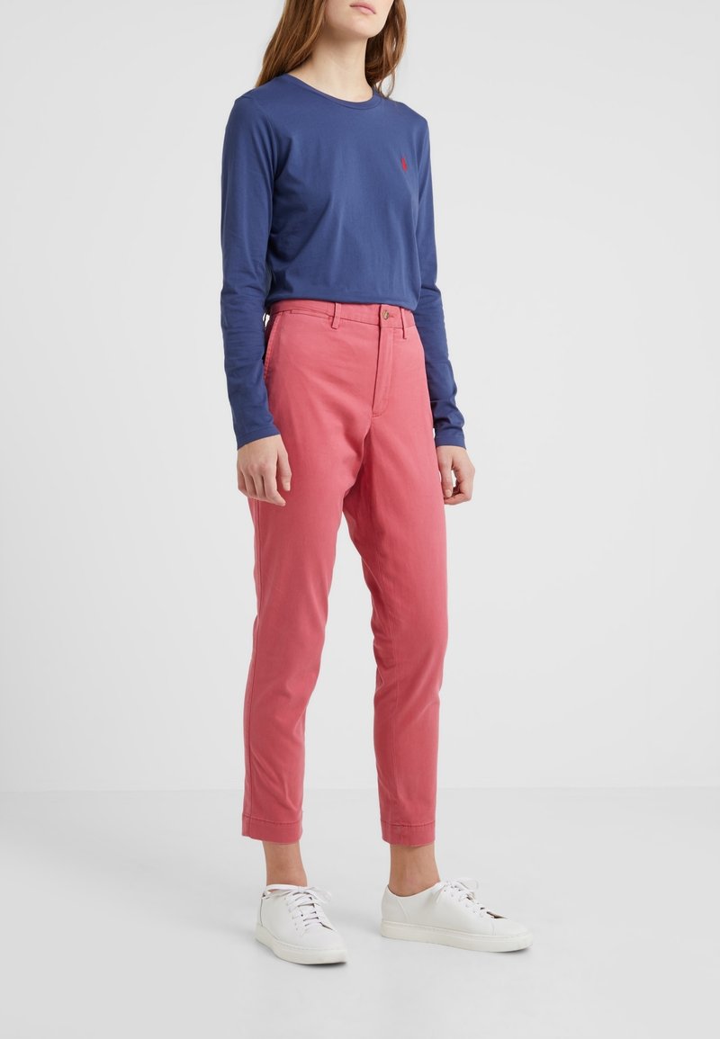 Personne portant une chemise bleue à manches longues, un pantalon rose taille haute et des baskets blanches, debout devant un fond clair uni.