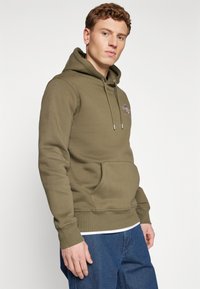 Felpa verde oliva con tasca a marsupio, cappuccio con coulisse e logo ricamato sul petto. Tessuto morbido con vestibilità comoda.