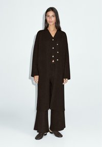 Long manteau en laine brun foncé avec des boutons dorés, porté sur un crop top assorti et un pantalon large. Le mannequin est posé sur un fond blanc.