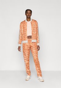 Oranžs leopardu raksta treniņtērps ar baltām svītrām, kas sastāv no bumbvedēja stila jakas un elastīgas jostasvietas biksēm, izgatavots no viegla auduma.