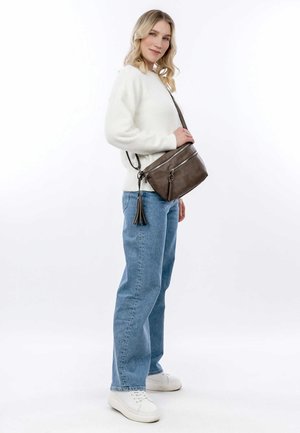 Brown leather crossbody bag with two zip compartments, a tassel accent, and a slender adjustable strap. Worn with a white sweater and blue jeans.