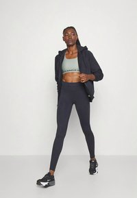Completo sportivo composto da un reggiseno sportivo verde chiaro, felpa con cappuccio nera con zip e leggings blu navy. Scarpe da ginnastica nere con dettagli bianchi.