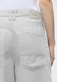Personne portant un pantalon gris clair avec de grandes poches arrière et une étiquette DRYKORN visible, debout devant un fond uni.