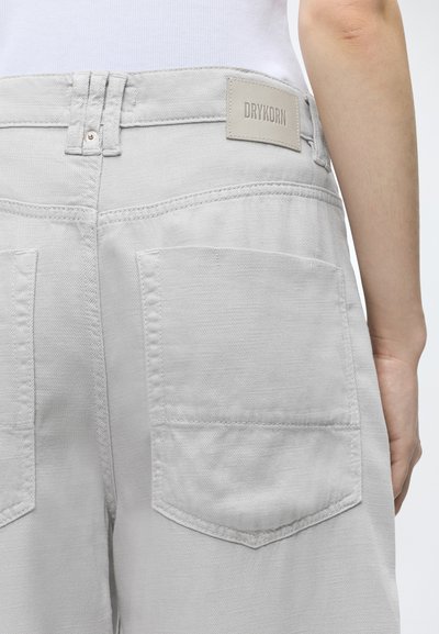 Personne portant un pantalon gris clair avec de grandes poches arrière et une étiquette DRYKORN visible, debout devant un fond uni.