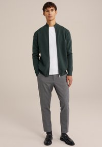 Tumši zaļš zips jaka ar augstu apkakli, uzvilkta virs baltas T-krekli, kombinēta ar gaiši pelēkiem biksēm un melniem uzvalka apaviem.