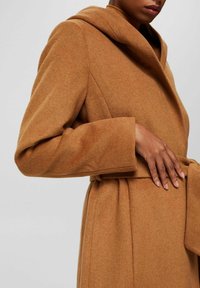 Vrouw in een camel kleurige wollen jas met een brede kraag en riem, hand op de taille rustend.