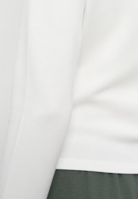 Gros plan d'une personne portant une chemise blanche à manches longues et un pantalon vert foncé, vue de côté et de la taille vers le bas.