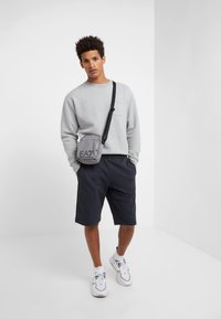 Felpa grigia, pantaloni corti neri e sneakers bianche. Piccola borsa a tracolla grigia con logo EA7. Look casual, texture semplici e design essenziale.