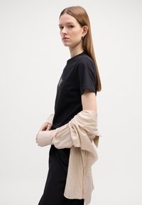 Sort t-shirt med frontdesign, paruet med en lys beige, struktureret, oversized, knap-op skjorte. Modellen poserer med armene over kors.