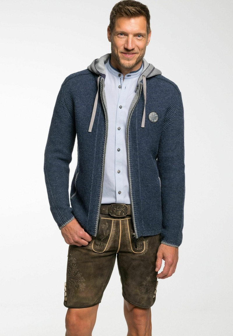 Marine gebreide cardigan met grijze capuchon, voorzakken en embleem. Gecombineerd met bruin leren shorts met fraaie stiksels en knopen.