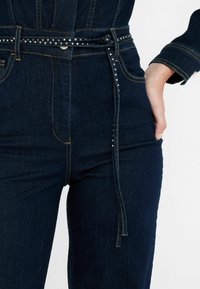 Pantaloni in denim blu scuro con vita alta, applicazioni decorative sulla cintura e cintura in tessuto annodata. Cuciture visibili in colore a contrasto.