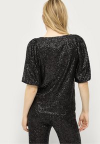 Haut noir à sequins avec de courtes manches bouffantes, surface texturée et encolure large. Le tissu reflète la lumière, créant un effet étincelant.