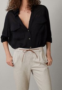 Schwarzes, locker sitzendes Button-up-Hemd mit zwei Brusttaschen, kombiniert mit hellbeigen Hosen, die einen braunen Kordelzug und seitliche Taschen haben.