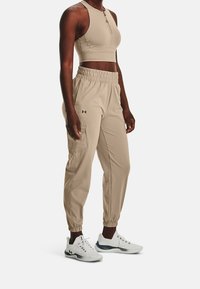 Žena v béžových sportovních teplákách s elastickým pasem a manžetami, doplněná o odpovídající zkrácený bezrukávový top a bílé sportovní boty.