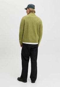 Oseba v olivno zelenem puloverju, črnih hlačah, črnih čevljih in temno zeleni kapici stoji obrnjen stran od preproste bele podlage.