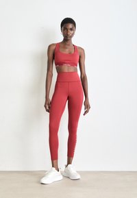 Vrouw staat in rode Calvin Klein sport-bh en high-waisted leggings met witte sneakers voor een effen muur achtergrond.