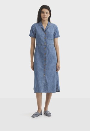 Femme portant une robe en denim bleu mi-longue à manches courtes et à boutons sur le devant, avec une poche avant, et des chaussures grises à enfiler, debout devant un fond uni.