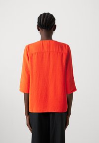 Camicia arancione, in tessuto testurizzato, con maniche a tre quarti e scollo rotondo. Presenta una cucitura posteriore e risvolti sulle maniche.