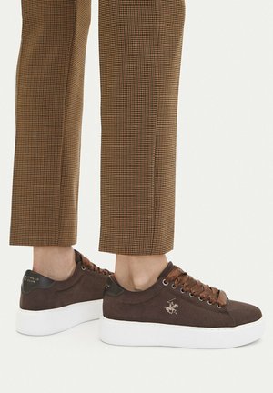 Bruna mockaplattformssneakers med satinsnören, använda med bruna rutiga byxor på vit bakgrund.