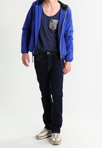 Blauer Jacke mit Reißverschluss und schwarzem Kragen, dunkelblaues Tanktop mit gemusteter Tasche, dunkle Jeans und beiger Sneaker mit weißen Sohlen.