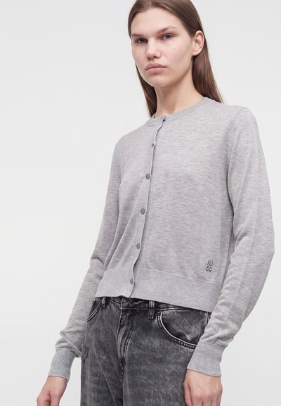 Jeune femme portant un cardigan gris clair boutonné et un jean noir délavé, debout devant un fond blanc uni.