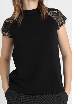 Femme portant un haut noir à col haut avec des manches courtes en dentelle florale, associé à un pantalon sombre à motifs, mains dans les poches.