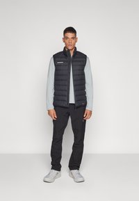 Schwarze Pufferweste mit hohem Kragen und horizontalen Nähten, getragen über einem hellblauen Langarmshirt, kombiniert mit schwarzen Hosen und grauen Schuhen.