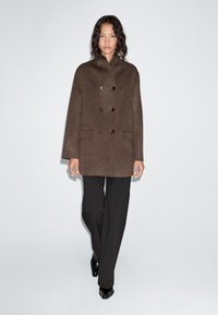Brown wool blend coat with a high collar, double-breasted design, two front pockets, and black buttons. Paired with black wide-leg trousers.