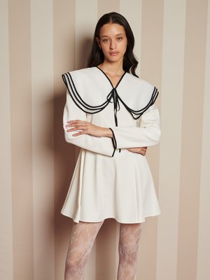 Vrouw met lang donker haar in een witte jurk met een grote marinierkraag met zwart stiksel en doorzichtige panty met bloemenpatroon, staande tegen een gestreepte muur.