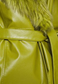 Gros plan d’un manteau en cuir vert avec une ceinture assortie et un col en fourrure texturé.