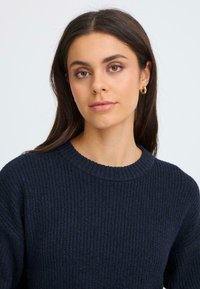 Sötétkék kötött pulóver bordás textúrával és kerek nyakkal, egy hosszú sötét hajú személyen bemutatva, arany karikafülbevalókkal.