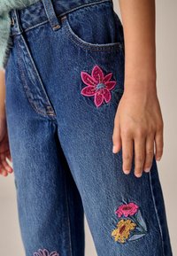 Jeans de mezclilla azules con diseños florales bordados en rosa, amarillo y verde. Corte recto, cintura alta y detalles de costura visibles.