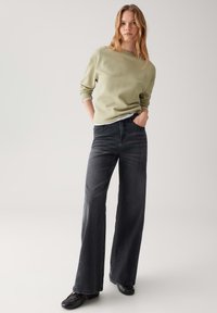 Giovane donna con capelli lunghi, che indossa un maglione verde chiaro a trama, jeans neri a gamba larga e scarpe nere, in piedi con le mani nelle tasche.
