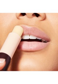 Trądzik do ust z zaokrąglonym końcem trzymany blisko miękkich, nawilżonych ust. Tube ma jasnoróżową bazę i ciemny akcent.